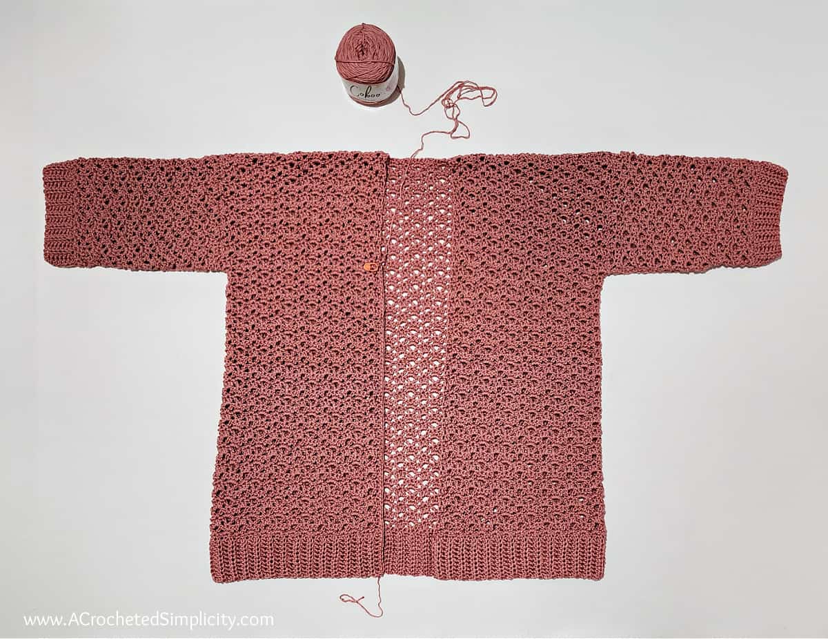 Mauve crochet summer cardigan laid flat on a white surface with a ball of leftover yarn.