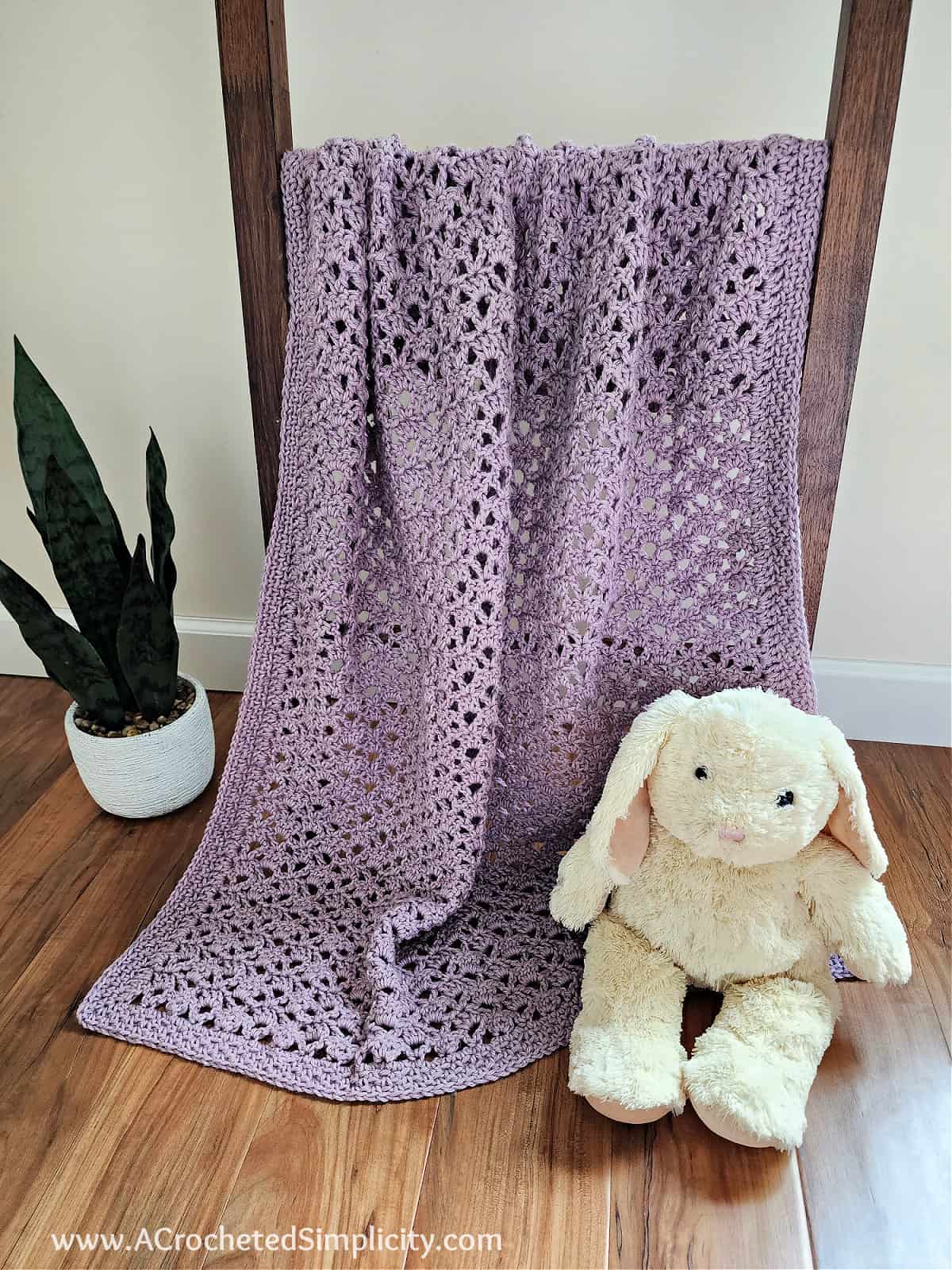 Lilac colored crochet lacy baby blanket with a cream colored stuffed bunny and plant