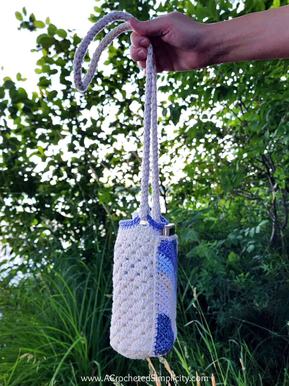 Someone holding a crochet water bottle holder in soft ecru and blue striped yarn with a phone pocket, showing the side view to see the granny stitch crochet.