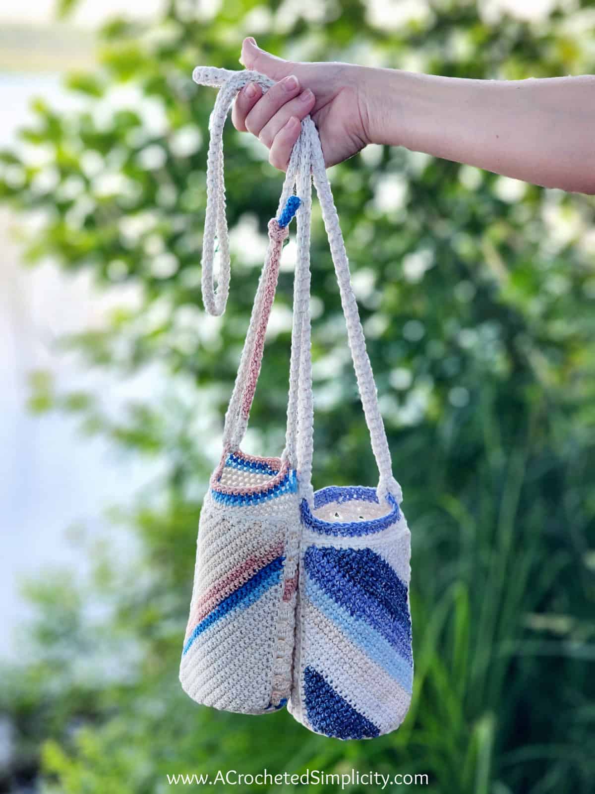 A woman's hand holding two crocheted water bottle holders in front of a green background by a lake.