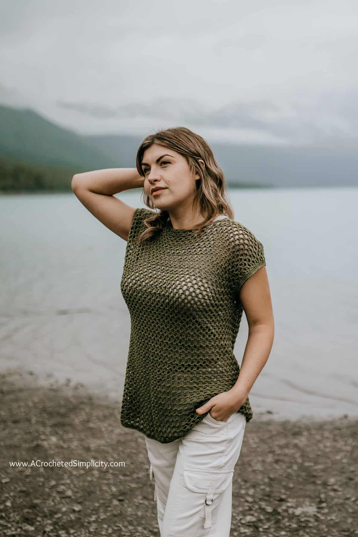 Woman modeling an olive crochet mesh top with mountains in the background.
