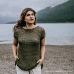 Model wearing a crochet mesh top with hands in her pockets while standing on a beach and looking off into the distance.