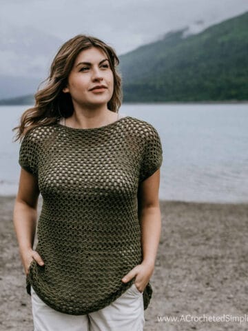 Model wearing a crochet mesh top with hands in her pockets while standing on a beach and looking off into the distance.