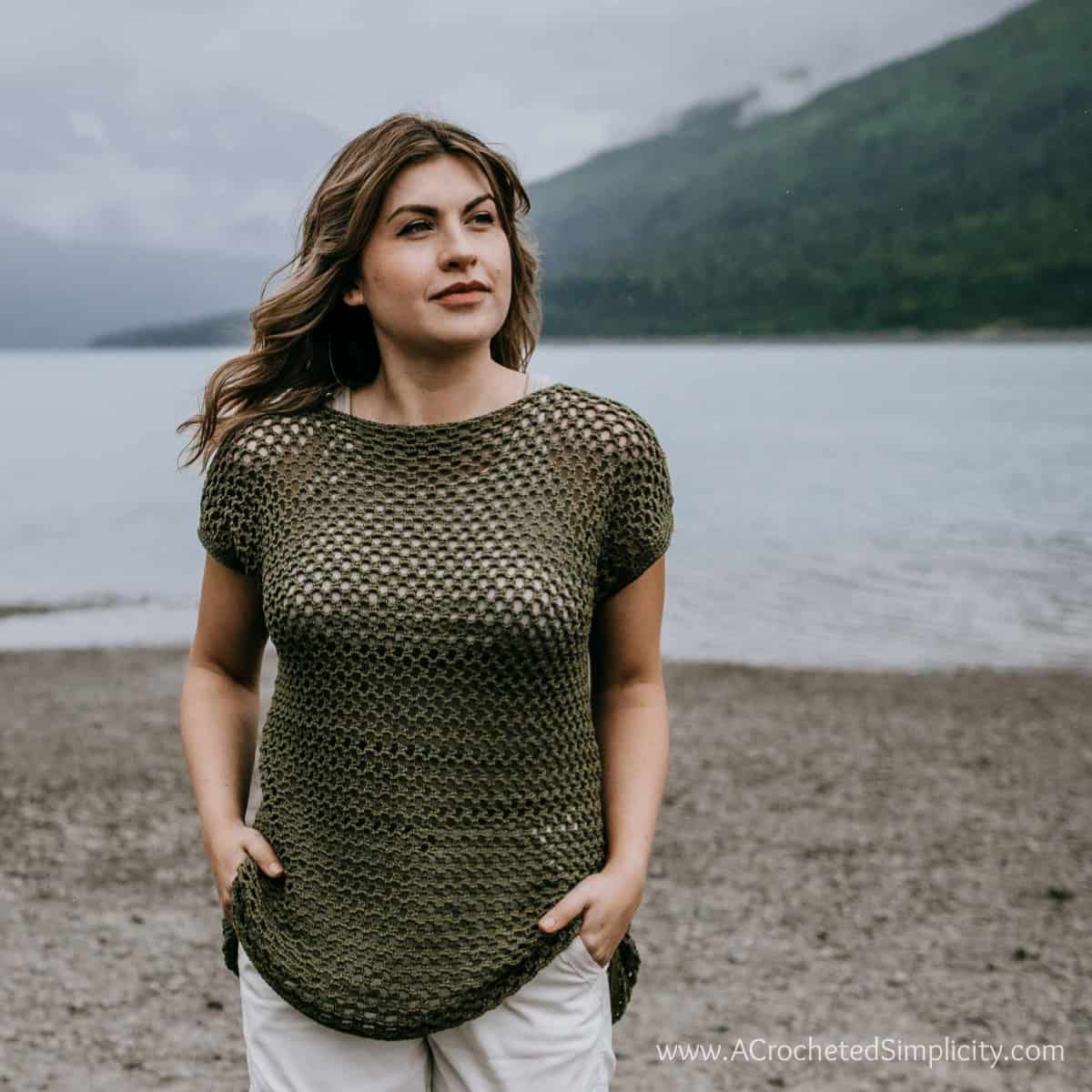 Model wearing a crochet mesh top with hands in her pockets while standing on a beach and looking off into the distance.