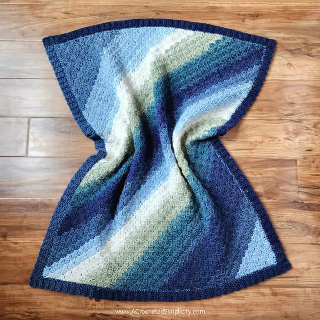 Diagonal box stitch C2C rectangle baby blanket in blue ombre yarn laying on a wood floor.