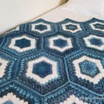 Crochet hexagon blanket in shades of blue and white yarn laying on white quilt.