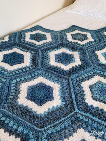 Crochet hexagon blanket in shades of blue and white yarn laying on white quilt.