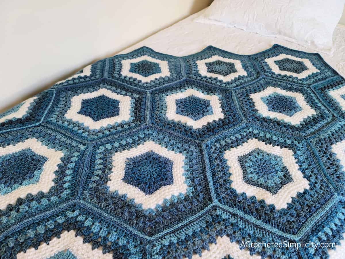 Crochet hexagon blanket in shades of blue and white yarn laying on white quilt.