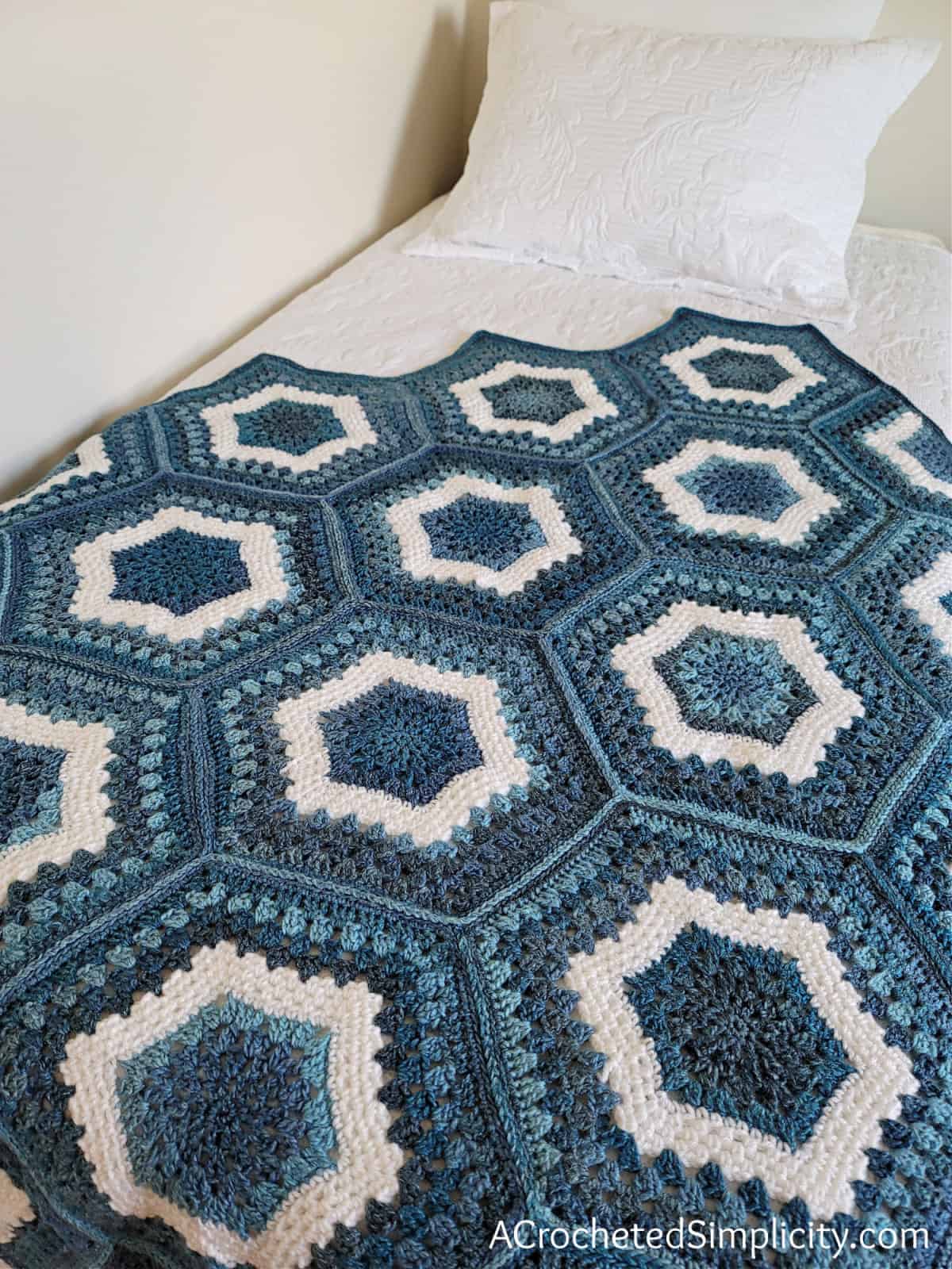 Hexagon crochet blanket in shades of blue and white yarn laying on white quilt.