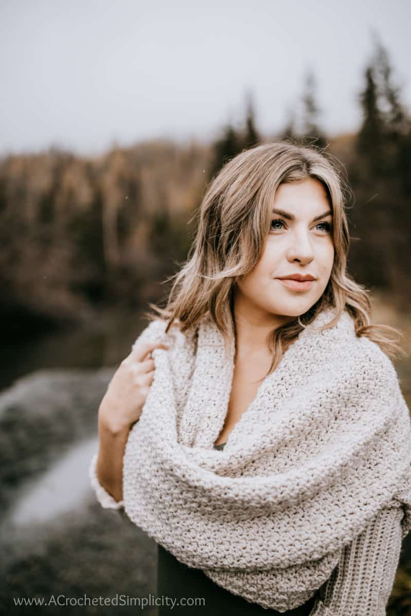 Woman modeling a crochet sweater scarf styled as a cowl outdoors near a river.
