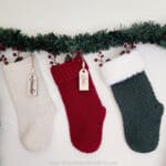 three crochet christmas stockings hanging with pine garland and berries