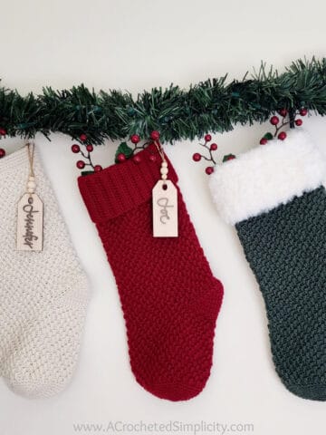 three crochet christmas stockings hanging with pine garland and berries