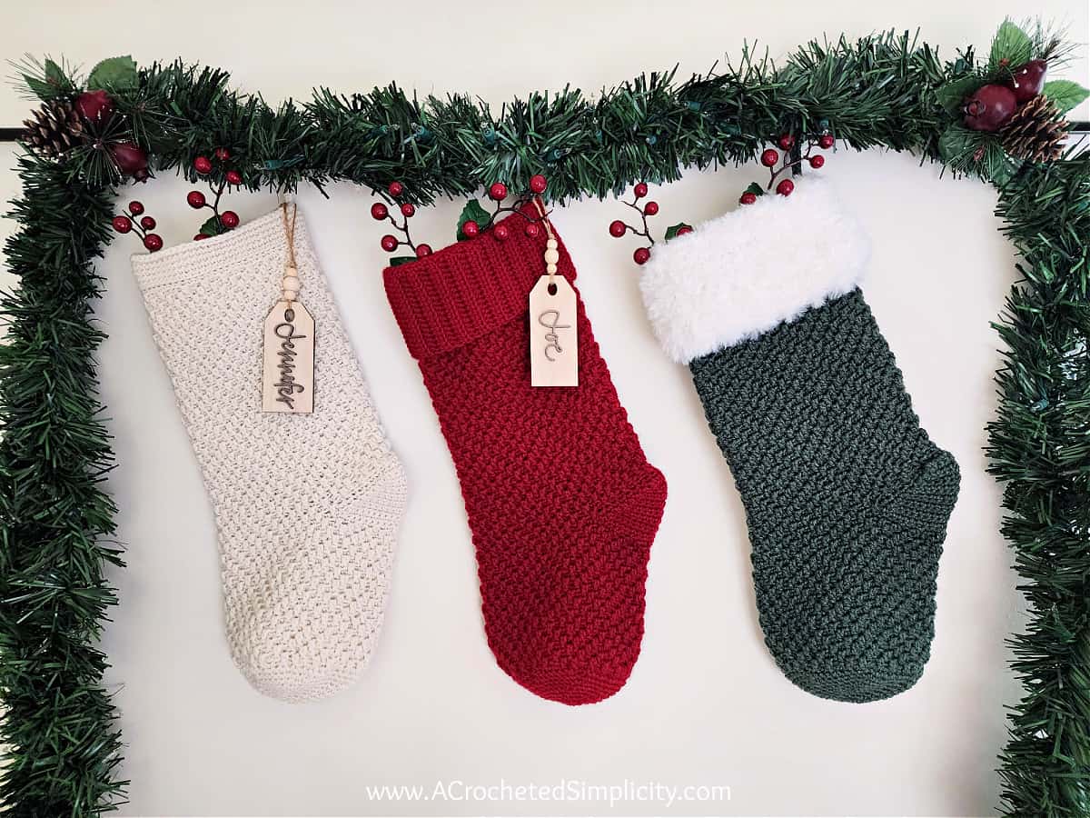 Three Christmas crochet stockings hanging with green garland and red berries