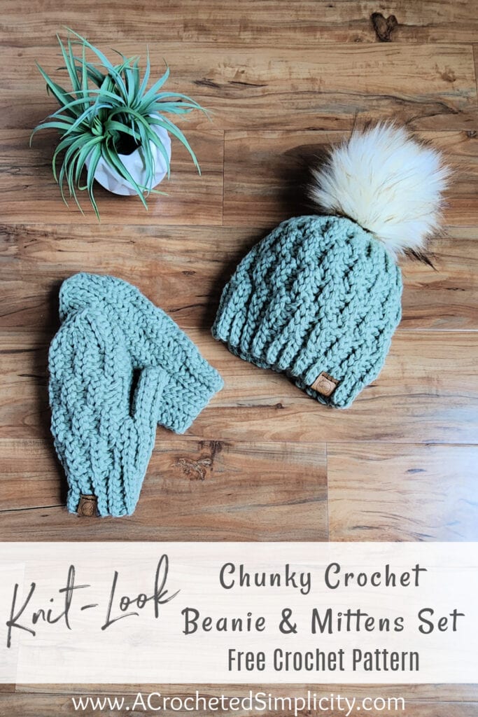 Chunky crochet beanie and mitten set with white faux fur pom laying on wood floor with small plant pinterest image