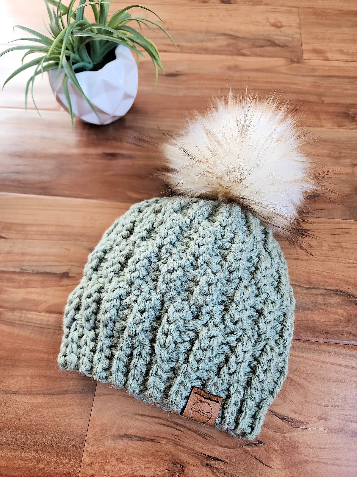 light green chunky crochet beanie with leather tag laying next to small potted plant