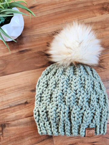 chunky knit look crochet beanie with white faux fur pom laying on wood with small white potted plant