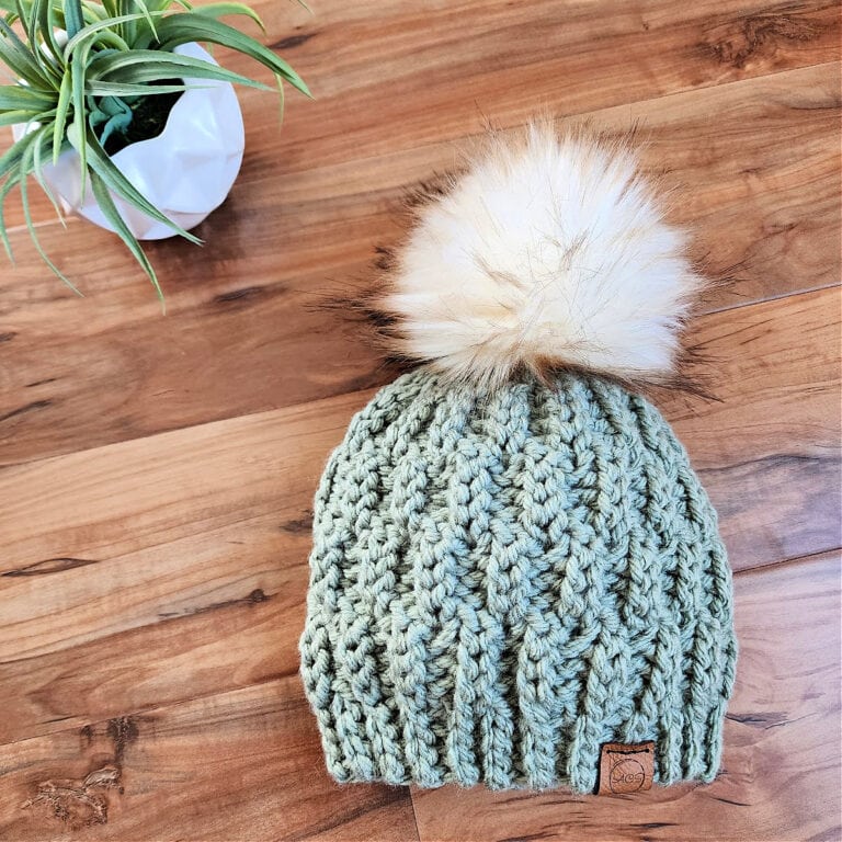 chunky knit look crochet beanie with white faux fur pom laying on wood with small white potted plant