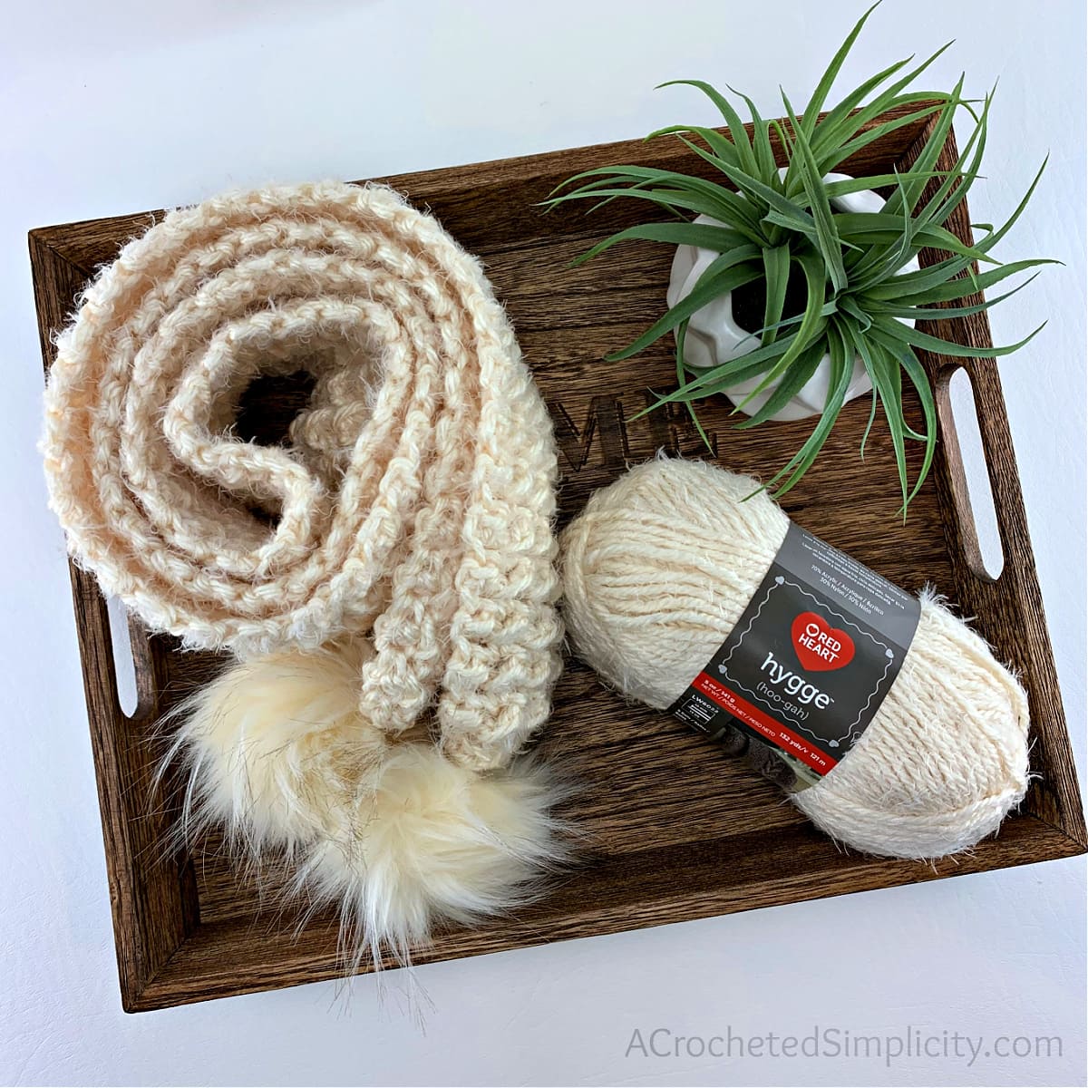 chunky crochet scarf in cream with faux fur poms rolled up on wooden tray with small plant