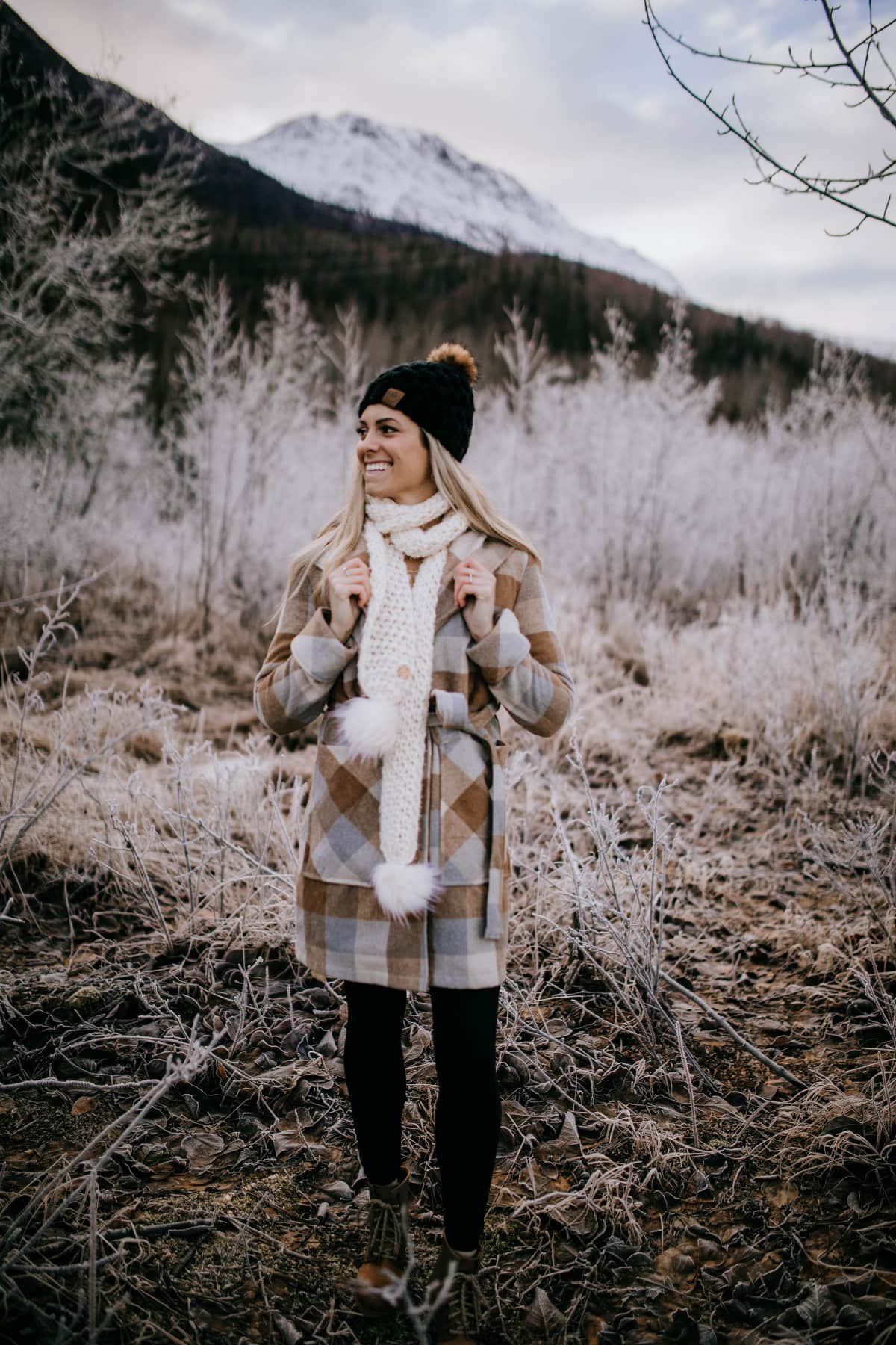 Woman modeling chunky crochet scarf in cream with faux fur poms in moutains