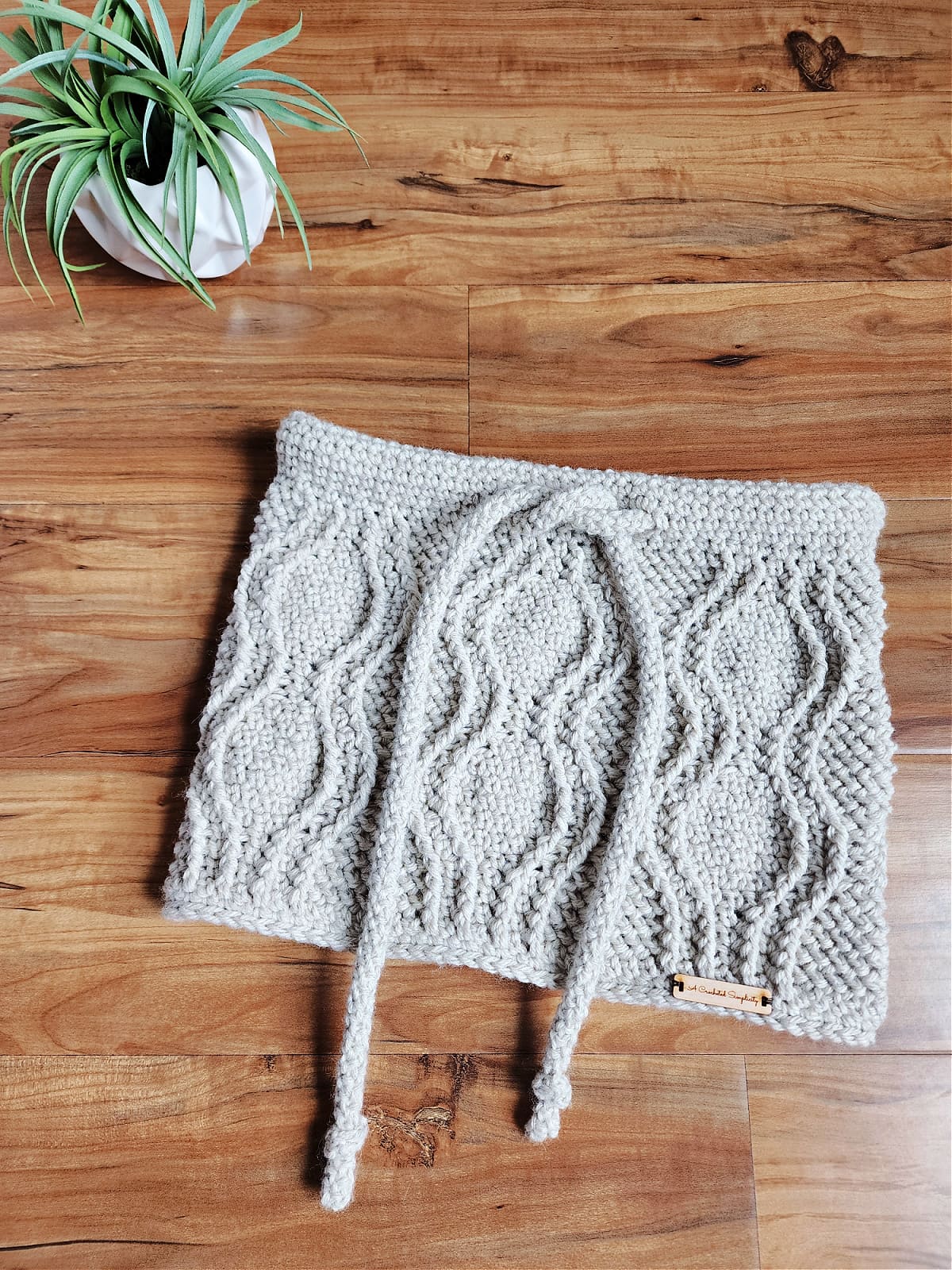 crochet cabled cowl laying on wood with small green plant