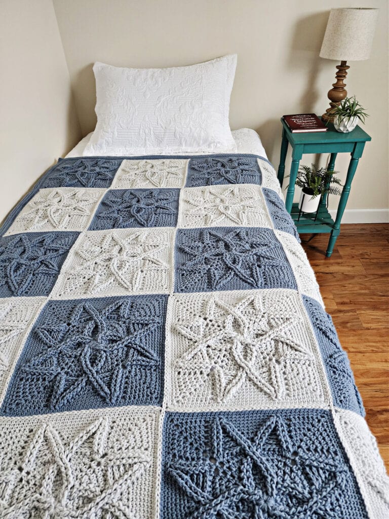 Crochet quilt with blue and grey crochet squares laying on guest bed near side table and small lamp.
