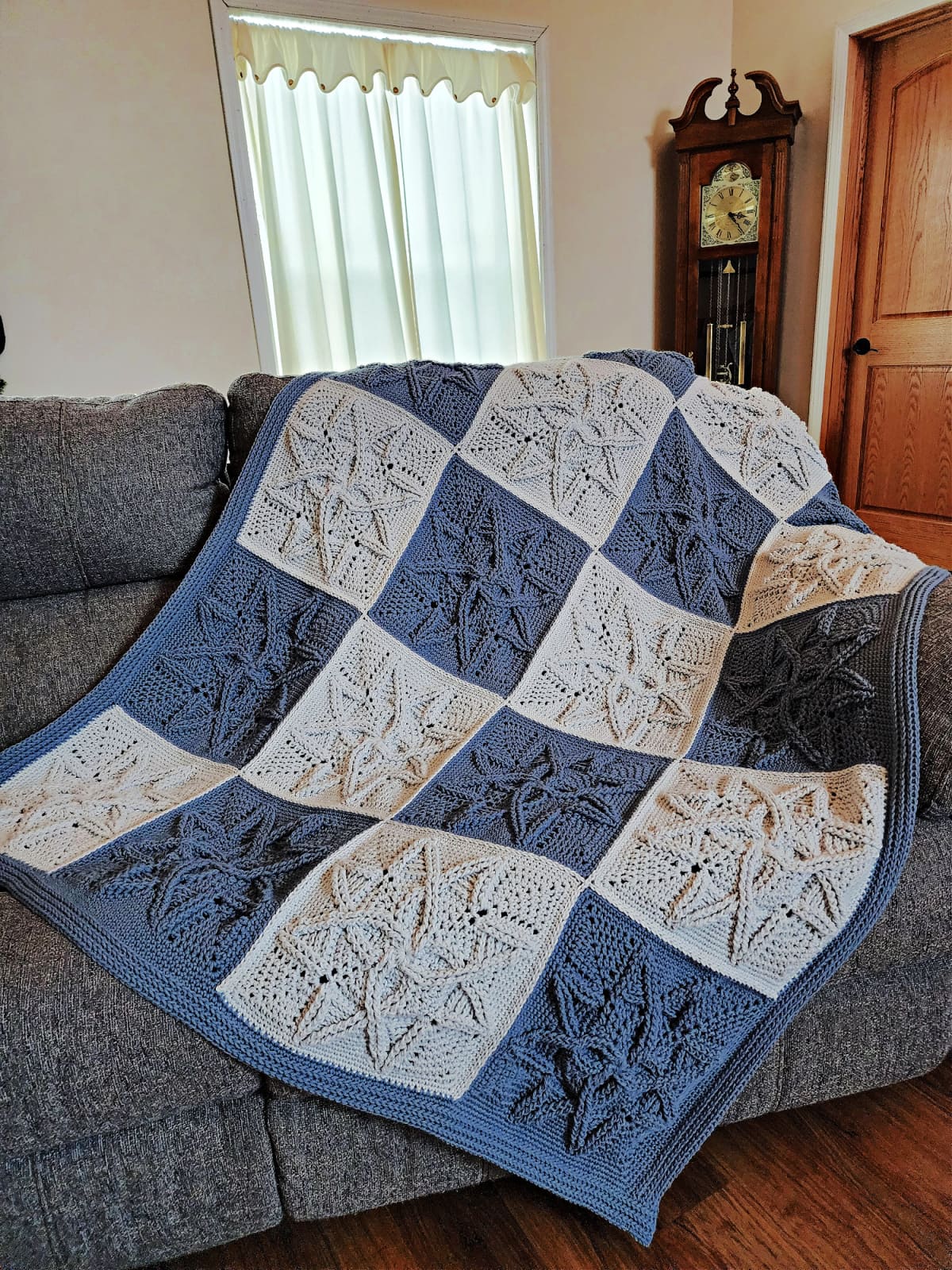 Cabled crochet blanket draped over a blueish-grey couch with grandfather clock in background.