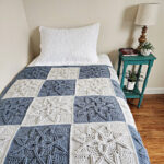 Steel blue and light grey cabled crochet blanket laying on small guest bed with side table and lamp.