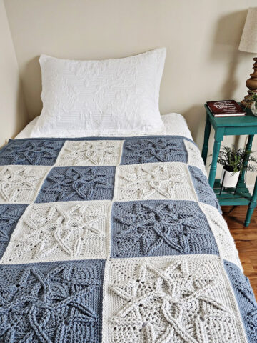 Steel blue and light grey cabled crochet blanket laying on small guest bed with side table and lamp.