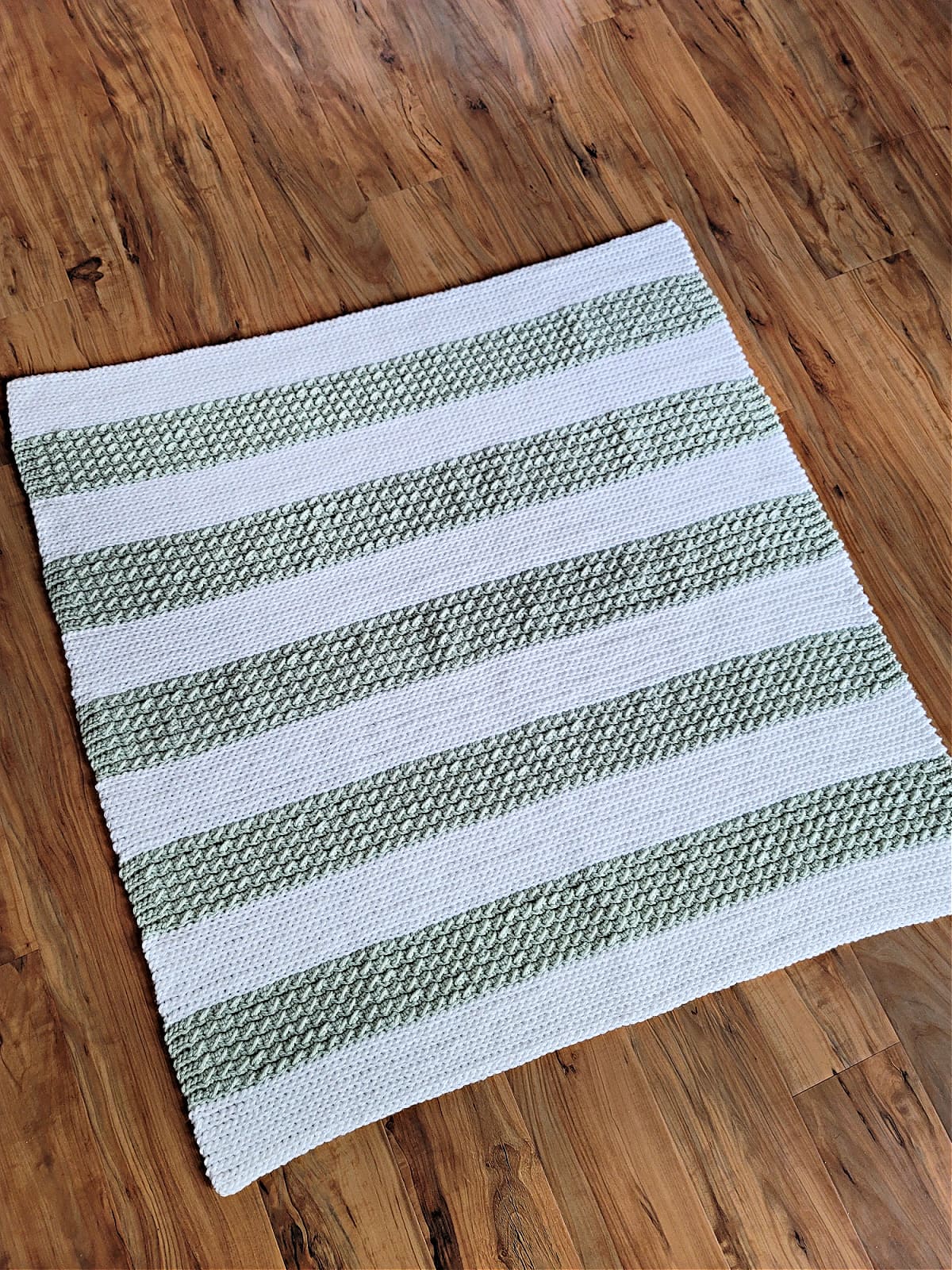 Crochet baby blanket pattern in white and greed wide stripes laid on wood floor.
