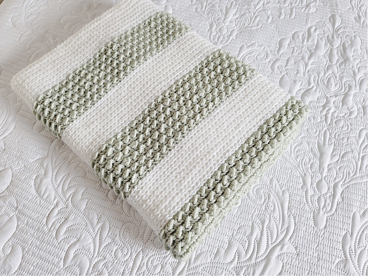 Striped crochet baby blanket in white and mint green folded and laying on white bedspread.