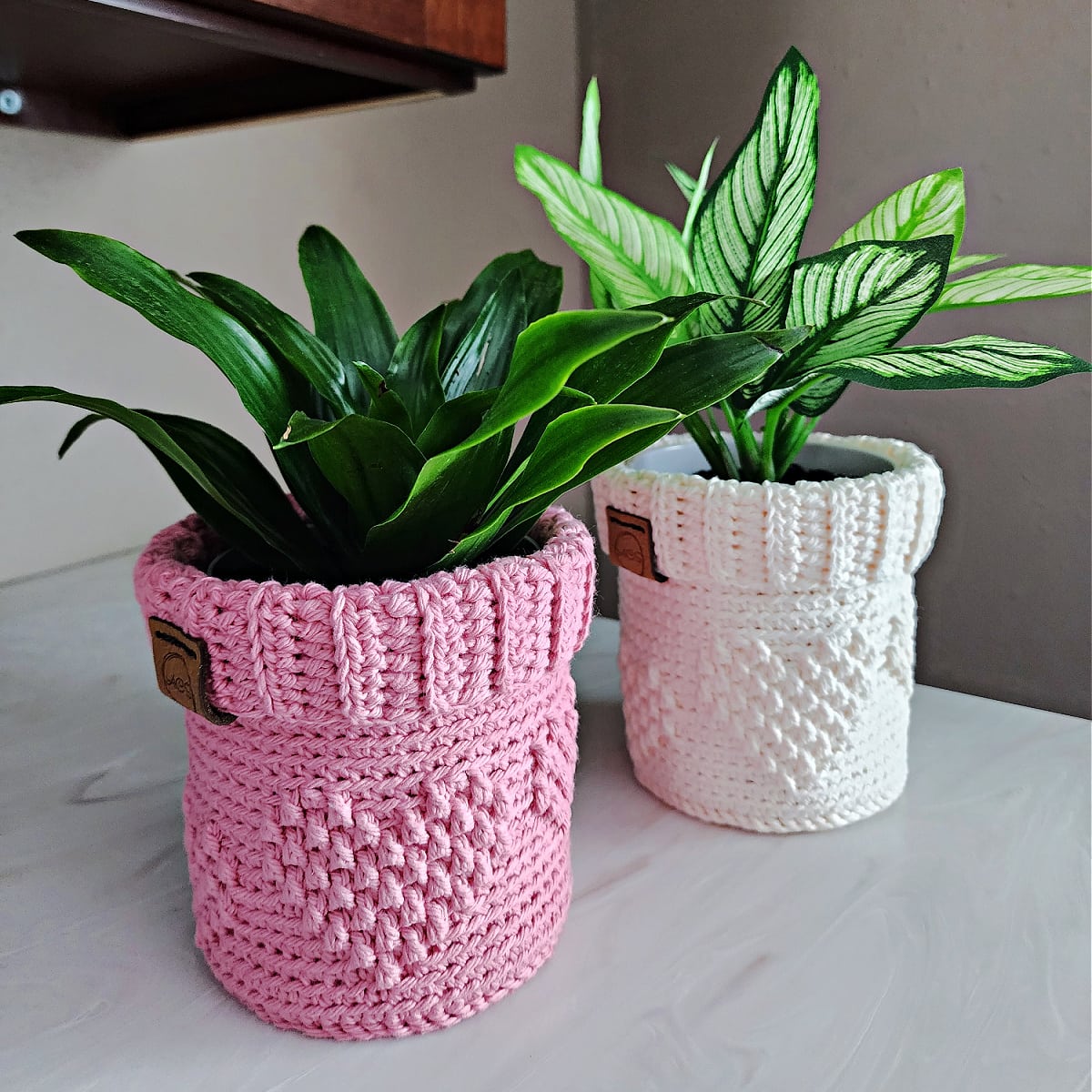 Pink and cream color crochet plant pot cover holding two indoor plants sitting on a marbled countertop.