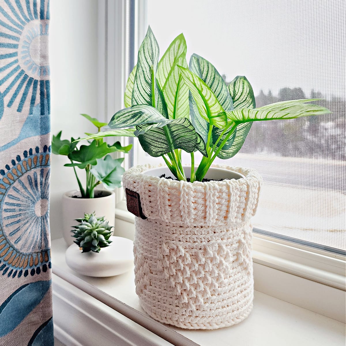 Cream color crochet plant pot cover with a potted plant sitting on a windowsill with blue curtain.