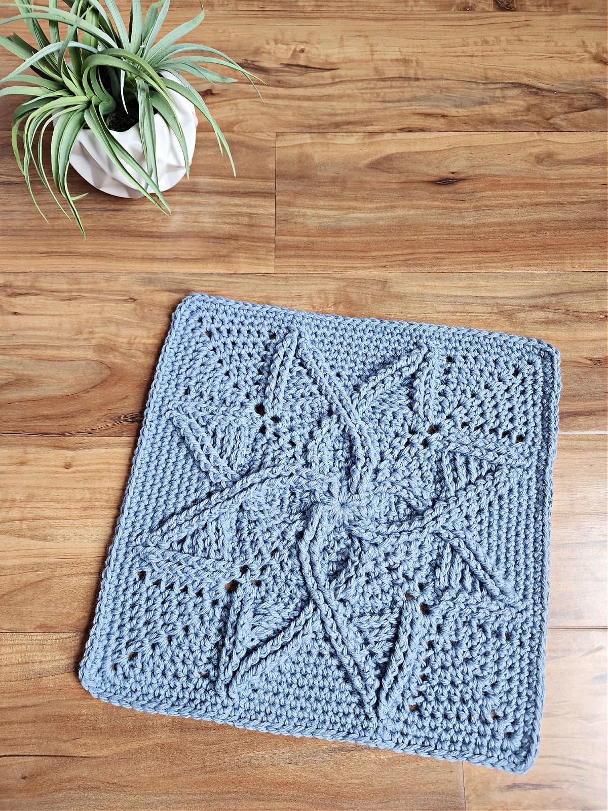Steel blue cabled crochet blanket square laying on wood floor with small potted plant.