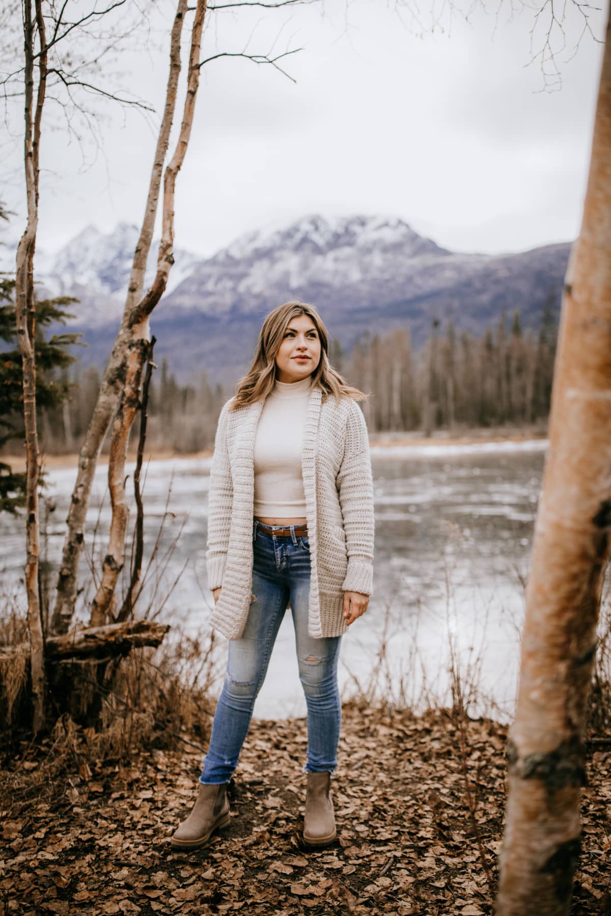 Woman modeling a light beige long crochet cardigan with wide ribbed collar near the mountains.