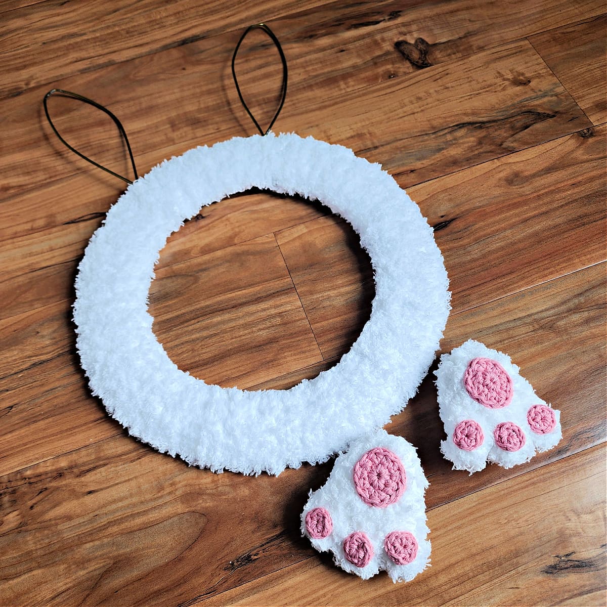 Two crochet bunny feet with pink toes and pads laying next to partially finished bunny wreath.