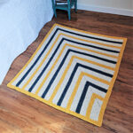 Side view of a chunky crochet blanket laying on a wood floor next to a bed.