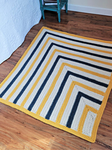 Side view of a chunky crochet blanket laying on a wood floor next to a bed.