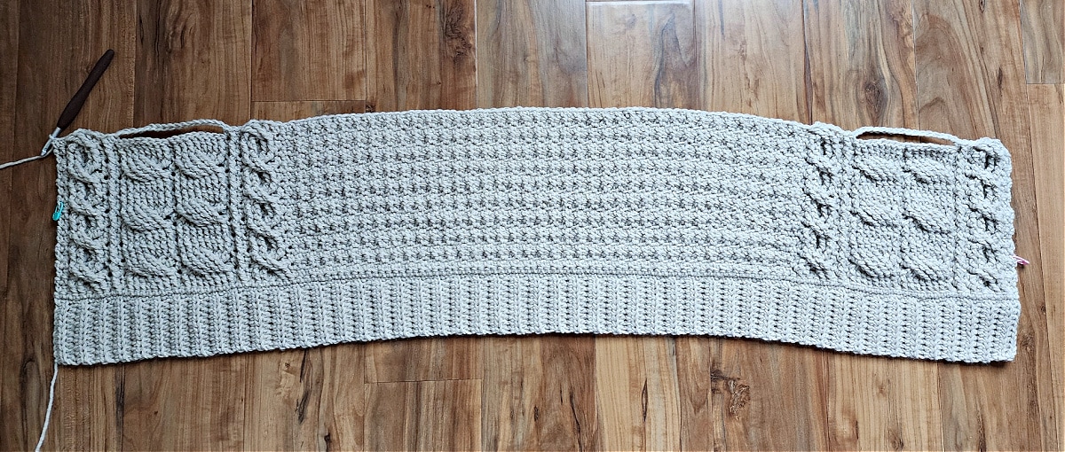 Beginning of crochet cable cardigan with pocket openings laying on wood floor.
