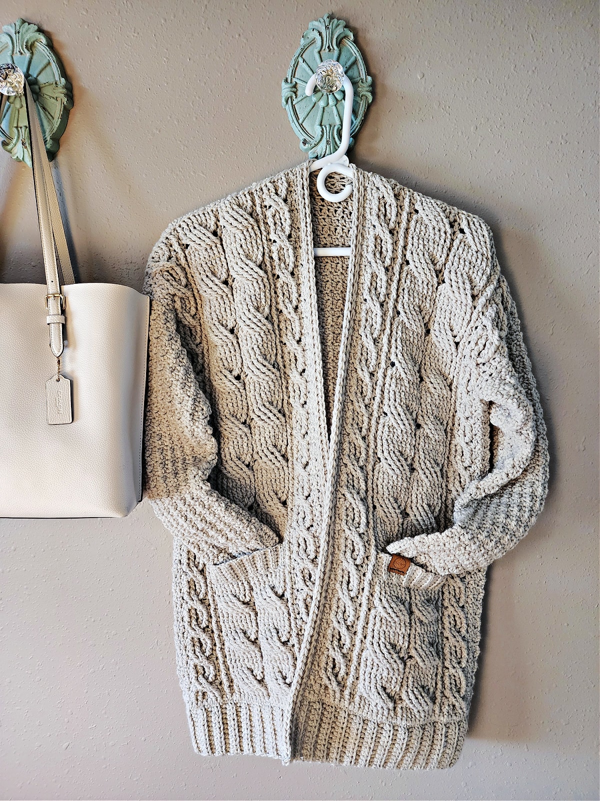 Light taupe crochet cable cardigan hanging on a hanger on a coat hook next to a white purse.