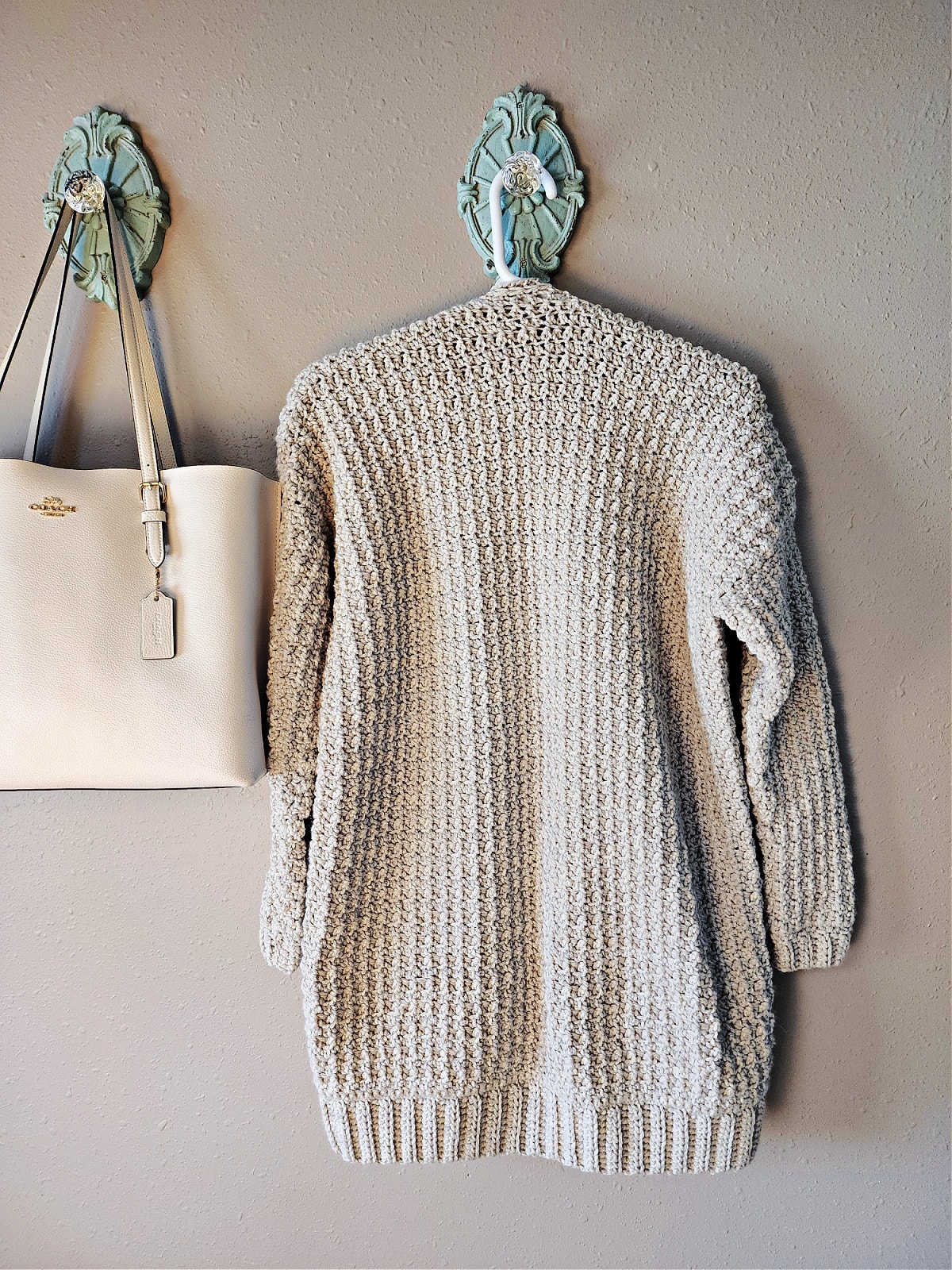 Back of cabled cardigan hanging on coat hanger next to purse.