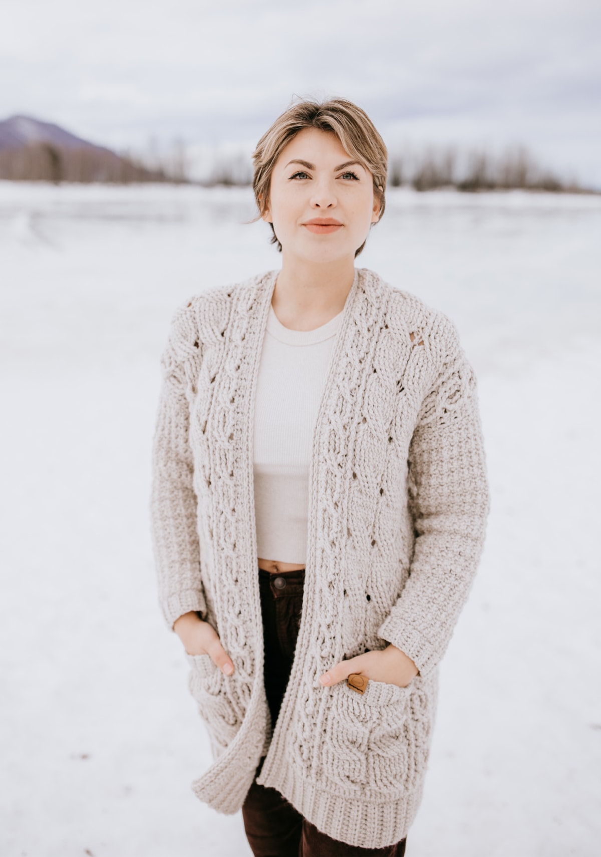 Woman with short hair modeling crochet cardigan near icy lake.