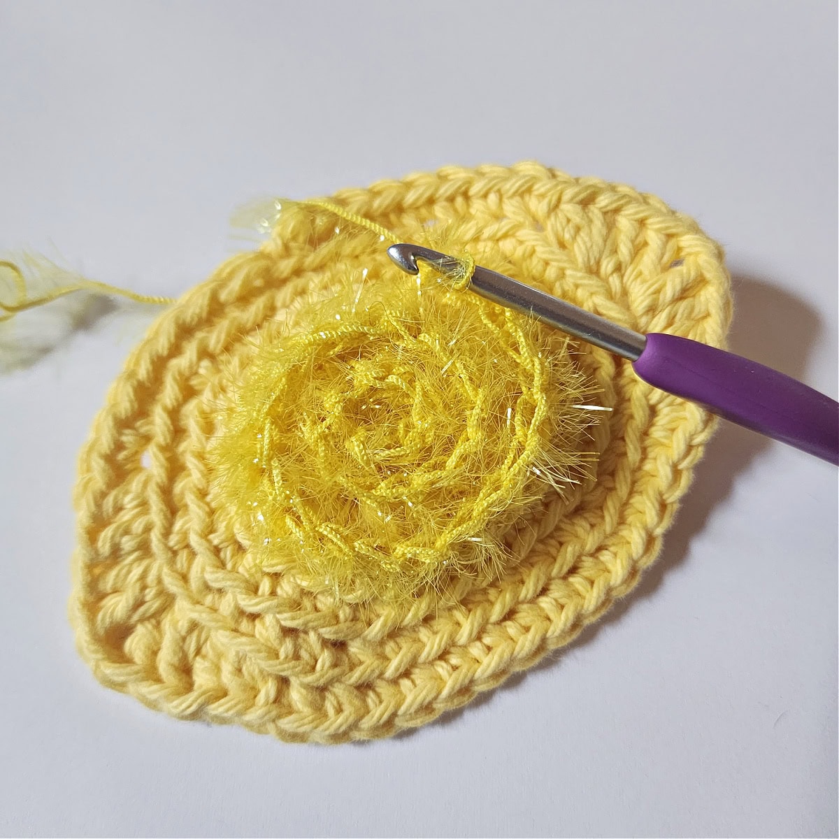 Crochet hook with scrubby sparkle yarn being added in a circle to a crochet lemon dish scrubby.