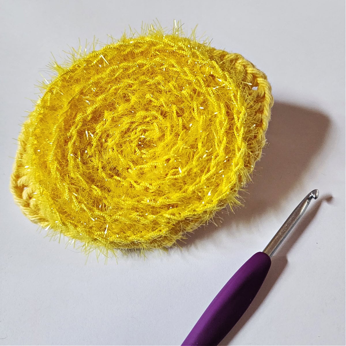 Crochet lemon with scrubby yarn added to the center of it and laying next to a purple 4.00 mm crochet hook.