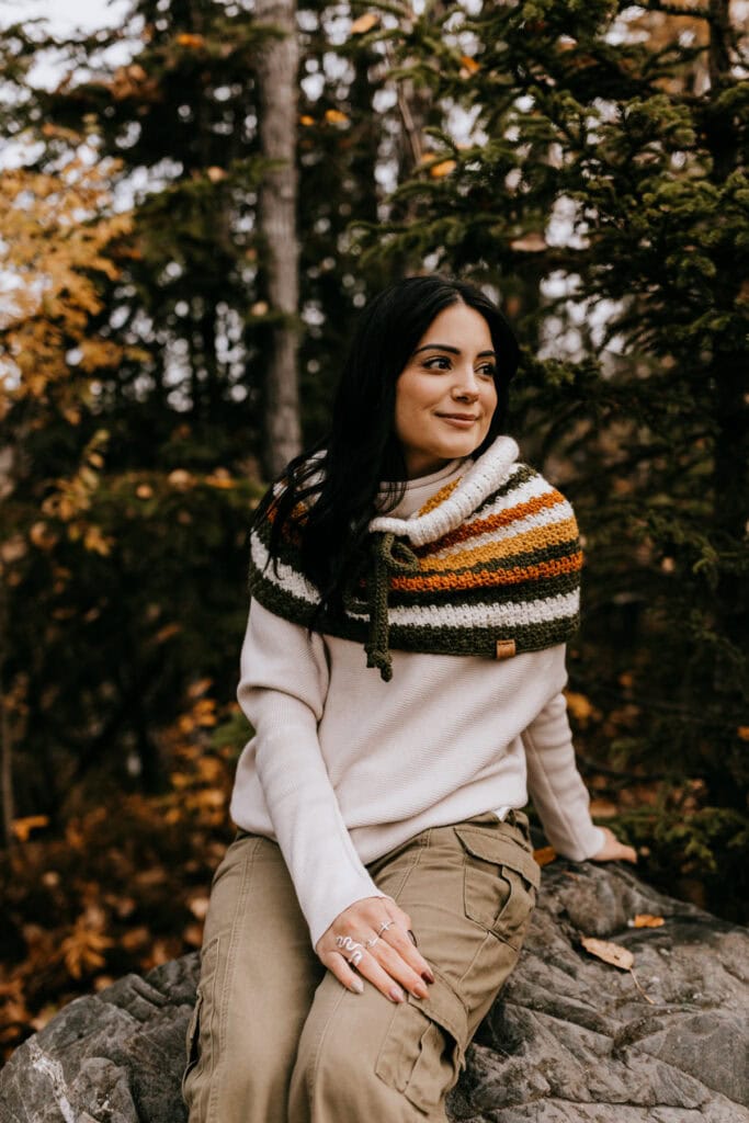 Young lady modeling a fall crochet cowl pattern while sitting on a rock with the cowl pulled down as a capelet.