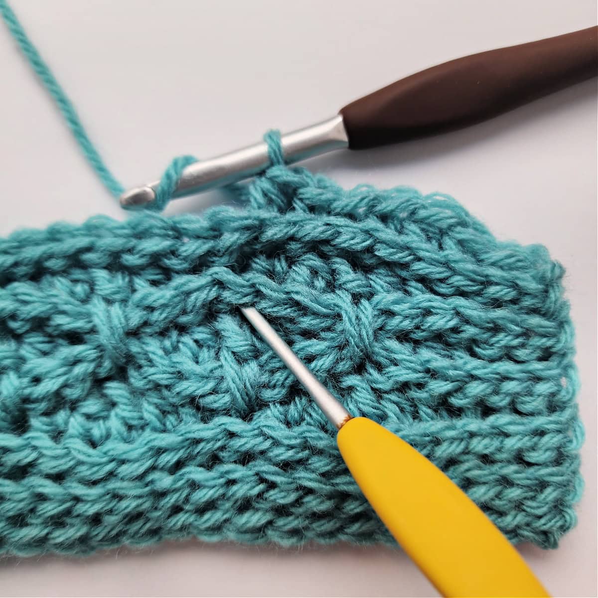 A yellow crochet hook showing where to work a double crochet stitch two rows below and a brown hook holding the working yarn.