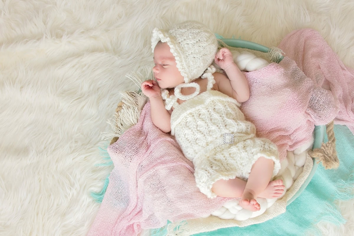 Baby in a basket wearing a white crochet romper and white bonnet.