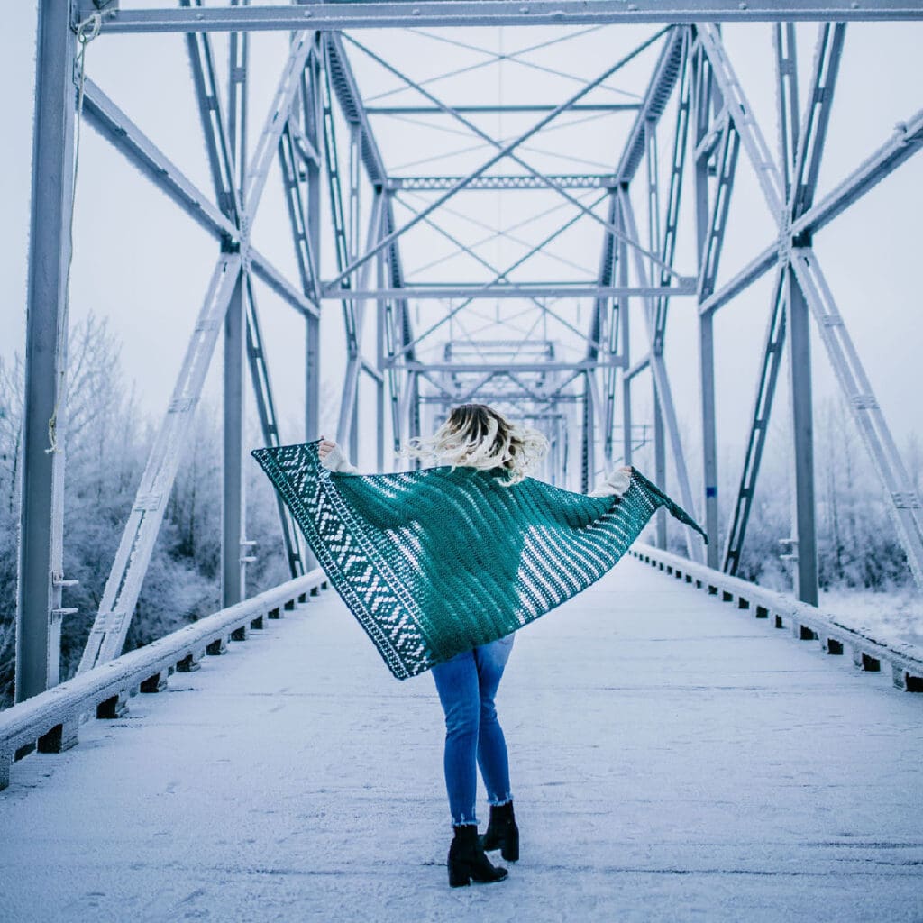 Woman standing on a bridge wearing blue jeans and a green crochet shawl.