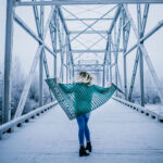 Woman standing on a bridge wearing blue jeans and a green crochet shawl.