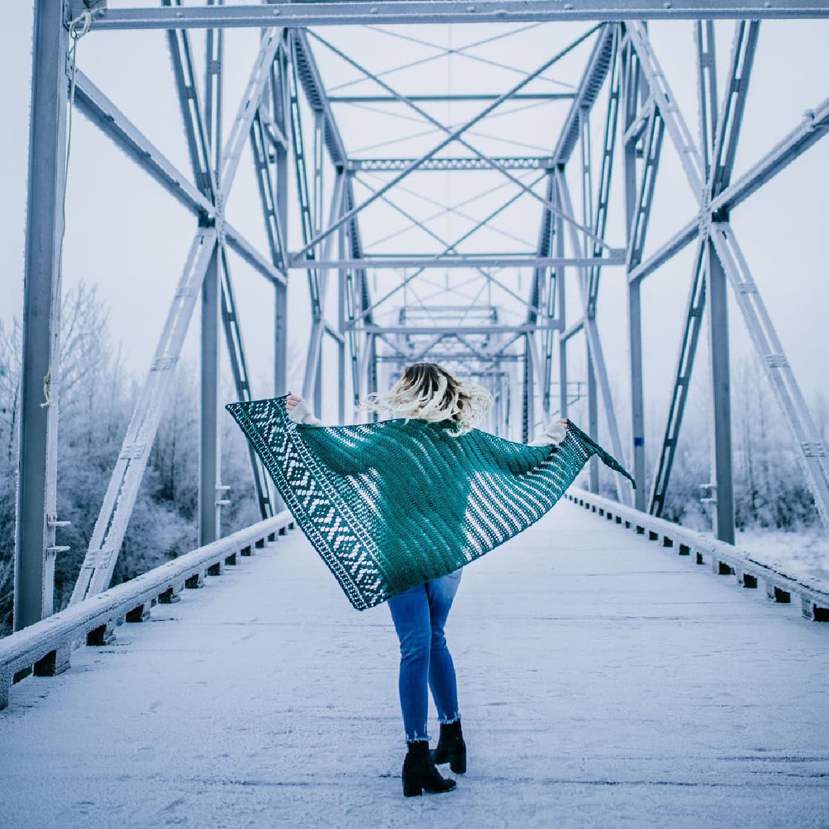 Woman standing on a bridge wearing blue jeans and a green crochet shawl.