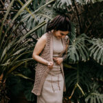 A young women wearing a long crochet vest over a knit dress standing in front of large greenery.